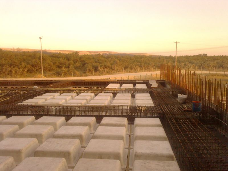 Şafak veya alacakaranlık vaktinde beton bloklar, donatı demetleri ve arka planda ağaçların bulunduğu bir inşaat şantiyesi manzarası